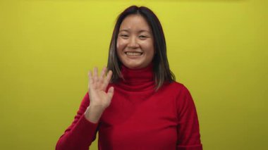 Kırmızı bir kazakla parlak sarı bir arka plana doğru mutlu bir şekilde el sallayan bir kadın izole edilmiş neşeli bir ortamda gençliğini ve pozitifliğini sergiliyor..