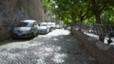 Roma 'nın eski kasabasındaki bulanık parke taşı caddesinde kaldırımda duran yeşil ağaçlar ve çözülmüş arabalarla Bokeh arka planı ve öğleden sonra ışıklarıyla sakin bir şehir manzarası yaratıyor..