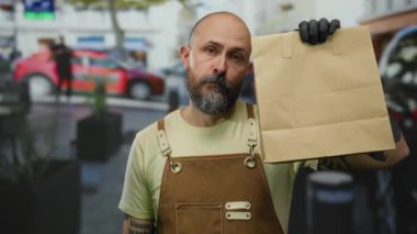 Şehirde sakallı bir adam, elinde kağıt torbayla önlük ve eldiven takıyor. Etrafı şehir çevresiyle çevrili..