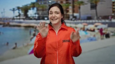 Kumsalda parlak turuncu hapishane tulumu giymiş bir kadın güneşli gökyüzünün altında neşeli bir ifadeyle el sallıyor..