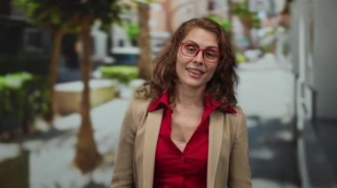 Kırmızı bluz ve bej ceket giyen, güneşli bir günde gözlüklü, açık havada gülümseyen, arka planda bulanık bir şehir manzarası olan, ifadesini gösteren bir kadın..