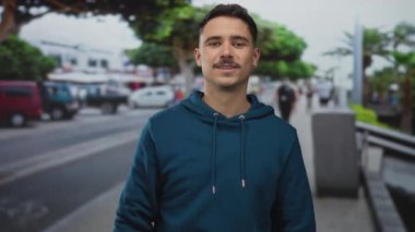 Mavi kapüşonlu genç adam güvenli bir şekilde gülümsüyor. Başparmakları şehir caddesini işaret ediyor. İçi araçlarla dolu ve insanlar canlı ağaçların önünden geçiyor..