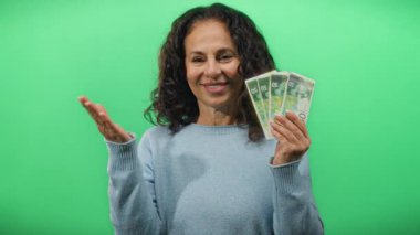 İsrail şekelleri tutan orta yaşlı bir kadın canlı yeşil bir arka plana gülümsüyor finansal ve izole bir ortamda paralarını sergiliyor..
