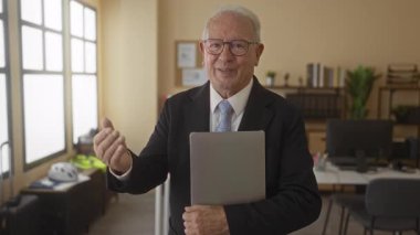 Ofis ortamında dizüstü bilgisayar tutan takım elbiseli kıdemli bir adam kendine güvenen bir ifadeyle profesyonel bir iş ortamı sergiliyor..