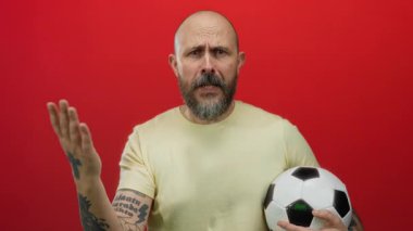 Sakallı bir adam futbol topunu canlı kırmızı arka planda tutuyor. Dövmeli elleriyle duygularını ifade ediyor..