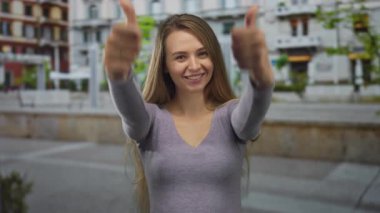 Mor gömlekli genç bir kadın arka planda renkli binalar olan bir şehir caddesinde baş parmak işareti yapıyor.