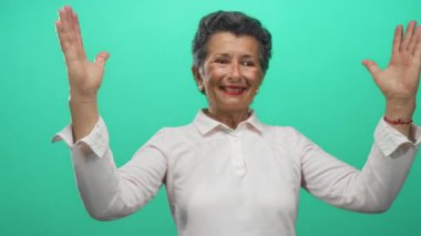 Yaşlı bir kadın, gri saçlı, iri yapılı, elleri parlak yeşil bir arka planı olan, mutluluk ve dışavurumluluk saçan..