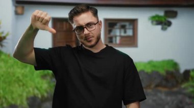 Sakallı genç bir adam şehir sokaklarında gözlüklü ve siyah tişörtlü, şehir ortamında karışık duygular ifade eden bir sokak sahnesinde başparmak kaldırıyor..