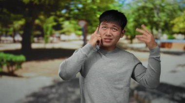 Güneşli bir günde parkta telefonla konuşan genç bir adam, yeşil doğayla çevrili jestlerle duygularını ifade ediyor..