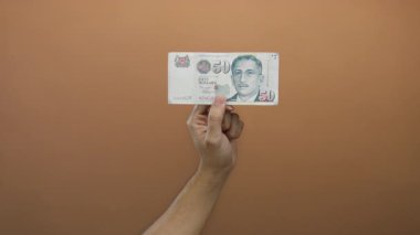 Kafkasyalı bir adam Singapur Doları banknotunu kahverengi duvarın arkasında tutuyor, soyutlanmış ve paranın detaylarına odaklanmış..