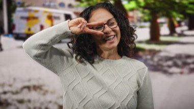 Şehir sokaklarında gülümseyen bir kadın kıvırcık saçlı ve gözlüklü neşeli bir jest yapıyor, rahat bir süveter giyiyor, neşeli bir orta yaşlı havası var ve şehir ortamında.