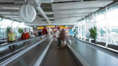 Bulanık insanlar, erkek ve kadınların olduğu havaalanı sahnesi. Terminalde yumuşak gün ışığı ve odaklanmış arka planda hareket halindeki bir yürüyüş yolunda..