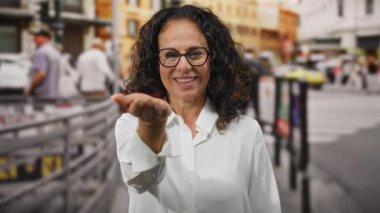 Beyaz tişörtlü, kıvırcık saçlı bir kadın şehir sokaklarında orta yaşlı, yetişkin İspanyol şehir ortamında jest yapmaya davet ediyor..