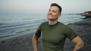 Gün batımında sakin bir plajda dururken güvenle gülümseyen dövmeleri olan genç bir adam. Okyanus dalgaları, berrak bir gökyüzünün altında yavaşça arka planda geziniyor..