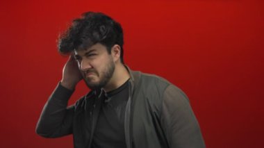 Sakallı genç İspanyol bir adam cesur bir kırmızı arka plana karşı kulak veriyor, merak ve konsantrasyon taşıyor..