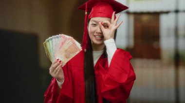 Mezuniyet elbisesi giymiş bir kadın, elinde Philippine Peso banknotları ile otelin içinde, kutlama yaparken neşeyle gülümsüyor..