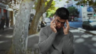 Gözlüklü genç adam şehir sokaklarında dikiliyor, telefonda konuşuyor, derin düşüncelere dalmış bir ifadeyle kentsel unsurlar ve doğal ışık ile çevrili..