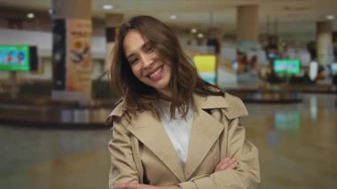 Bej ceketli genç bir kadın kapalı havaalanı terminalinde duruyor. Güvenle gülümsüyor. Arkaplan dolusu bulanık reklam ve ışığa karşı kollarını kavuşturmuş.