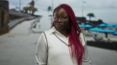 Sahilde kırmızı örgülü Afro-Amerikalı bir kadın, neşeli bir dille, kumsalda güneşli bir günün tadını çıkarıyor parlak şemsiyeler ve sakin bir arka planla.