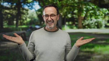Gündelik kıyafetli İspanyol bir adam, yemyeşil ağaçların altında sergilenen sakin bir parkta avuçlarını açıyor, düşünceli ve boş zamanlarını dışarıda geçiriyor..