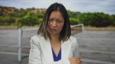 Beyaz ceketli bir kadın tenis kortunun dışında duruyor. Etrafı yeşilliklerle çevrili düşünceli bir ifadeyle sakin ama içsel bir anın altını çiziyor..