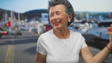 Gri saçlı yaşlı kadın, kalabalık bir sahil limanında tekne ve denizle gitar çalarken neşeli bir şekilde taklit ediyor, açık havada canlı bir müzikal ifade anı sergiliyor..