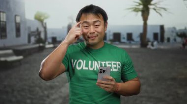 Açık havada güneşli bir günde deniz kenarındaki bir gezinti alanında elinde akıllı bir telefonla gülümseyen yeşil gönüllü tişörtü giyen genç bir adam..