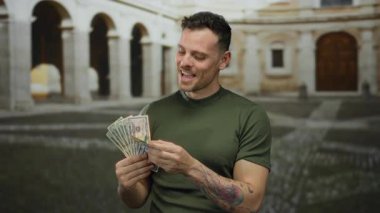 Açık üniversite ortamında Amerikan doları tutan genç bir adam, eski bir üniversite avlusunda renkli dövmeler sergilerken mutluluk ve zenginlik sergiliyor..