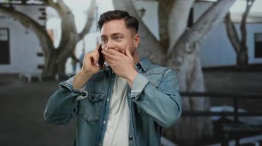 Kot ceketli genç adam parkta telefonla konuşuyor. Arka planda beyaz binalar ve ağaçlar bulanık. Mutluluğu ifade ediyor..