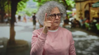 Gri saçlı yaşlı kadın şehir sokaklarında ihtiyatlı bir jest yapıyor, şehir manzarasının ortasında dikkat çekiyor..