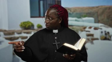 Gözlüklü ve papaz kıyafeti giyen kırmızı örgülü bir kadın kitabı ve puanları elinde tutarken açık havada, yeşil ve taş basamaklı binaların yanında duruyor..