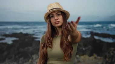 Deniz kıyısında jest yapmaya davet eden kadın, sakin okyanus manzarasına tepeden bakan ciddi bir ifadeyle hasır şapka takıyor.
