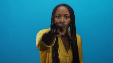 Mavi bir duvarın önündeki odak noktasını işaret eden kadın, sarı bir gömlek giyiyor, özgüven ve yoğunluk yayıyor, jest ve ifadeyi vurguluyor..