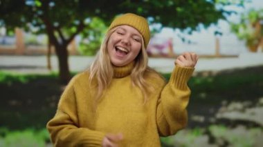 Sarı kazaklı ve bereli bir kadın, güneşli bir günde yeşil ağaçların arasında gitar çalıp eğleniyormuş gibi yapıyor..