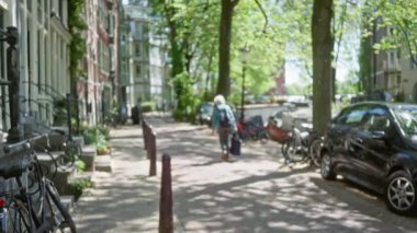 Hollanda, Amsterdam 'da ağaçlarla kaplı bir caddede park halindeki bisiklet ve arabalarla yürüyen bir kadının görüntüsü bulanık..