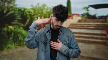 Kot ceketli bir adam şehir caddesinde duruyor, giysileri ayarlıyor, arka planda yeşil yapraklar var, rahat bir şehir atmosferiyle çekici bir açık hava ortamı yaratıyor..