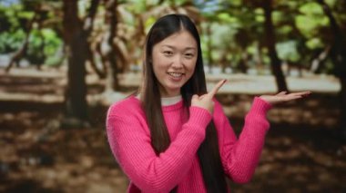 Pembe kazaklı genç bir kadın, güneşli bir park ortamında gülüyor, yemyeşil ağaçlarla çevrili, neşeli ve rahat bir açık hava sergiliyor..