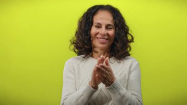 Kıvırcık saçlı olgun bir kadın neşeli bir şekilde canlı sarı bir arka plana alkışlıyor, izole bir ortamda mutluluk ve pozitiflik yayıyor..