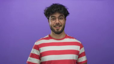 Sakallı ve gözlüklü genç adam kibarca gülümsüyor ve dudaklarını parlak mor bir arka planla örtüyor. Kırmızı çizgili gömleğiyle, eğlence ve samimi bir duyguyu çağrıştırıyor..