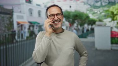 Açık şehir ortamında telefonla konuşurken gülümseyen olgun bir adam. Arka planda yeşillik ve gündüz çekilen binalar var.