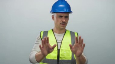 Yansıtıcı yelek ve şapka giyen İspanyol genç adam, tecrit edilmiş beyaz bir arka plana karşı reddedici bir jest yapıyor. Tereddüt veya ihtiyatı dile getiriyor..