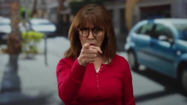 Kırmızı gömlekli son sınıf İspanyol kadın şehir sokaklarında dikiliyor, şehir arabaları ve arka planında ciddi bir ifadeyle parmakla işaret ediyor..