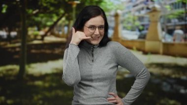 Ağaçlar ve bulanık şehir geçmişiyle parkta el kol hareketi yapan, gözlük takan ve gri kazak giyen, şakacı, kendine güvenen vücut diliyle açık havada mizahını ifade eden bir kadın..