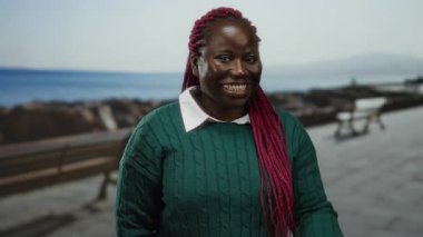 Kırmızı örgülü bir kadın açık bir günde deniz kenarında gezinirken gülümseyerek küçük bir jest yapar..