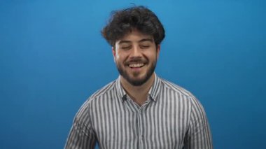 Sakallı genç adam çizgili gömleğiyle canlı mavi bir arka plana karşı güvenle gülümsüyor, gözleri kapalı ve elleri göğsünde mutluluk ve rahatlama yayıyor.