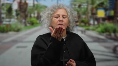 Gri saçlı ve rahip kıyafetli yaşlı bir kadın bir şehir caddesinde duruyor, elinde bir haç, modern bir şehirde maneviyatı ve dini bağlılığı simgeliyor..