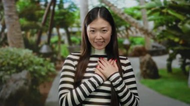 Kadın açık hava parkında duruyor, elleri göğsünde, çizgili tişört giyiyor, yemyeşil, sıcacık gülümsüyor, huzurlu, huzurlu bir atmosfer yaratıyor..