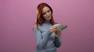 Elinde İsviçre banknotları tutan kadın, izole edilmiş pembe bir arka plana gülümsüyor. Kırmızı saçlarına ve güzel ifadesine odaklanmış para ve finansal konseptleri sergiliyor..