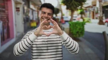 Genç adam elleriyle kalp şekli yaratıyor, canlı bir şehir caddesinde sıcacık gülümsüyor, canlı şehir havasını yakalıyor.