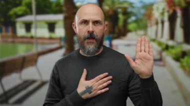 Parkta el pençe divan duran, kel ve sakallı, gri bir kazak giyen ve samimiyetini gösteren bir adam..